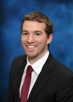 Man in white shirt, red tie, and black blazer, against a blue background